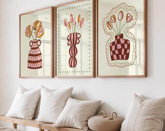Puede incluir: Tres impresiones artísticas enmarcadas con arreglos florales en jarrones decorativos. Los jarrones tienen patrones y formas únicas, y las flores son en tonos naranja, rosa y amarillo. Los marcos son de un cálido tono de madera, sobre un fondo crema neutro.