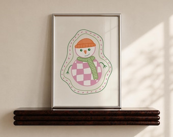 May include: A framed print featuring a stylised snowman illustration with an orange beanie, a green striped scarf, and a pink and white checkered body, all enclosed within a wavy green outline on a white background.