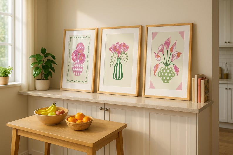 May include: Three framed floral art prints with wooden frames. Each print features a different arrangement of pink flowers in a vase. The prints are displayed on a white cabinet with a wooden table in front, holding fruit.