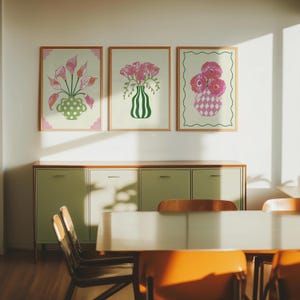 以下が含まれることがあります： 3つの額入りフラワーアートプリント。各プリントには、装飾的な花瓶に入ったピンクの花が描かれています。花瓶は緑色で、フレームは明るい木の色です。プリントは、明るい緑色のキャビネットの上に掛けられています。