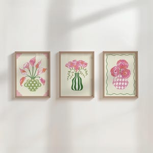 以下が含まれることがあります： 3つの額入りアートプリントのセット。花瓶に活けられた花の絵柄です。各プリントは、ピンク、緑、クリーム色を基調とした異なる花瓶のデザインとアレンジメントを特徴としています。フレームは天然木の色です。