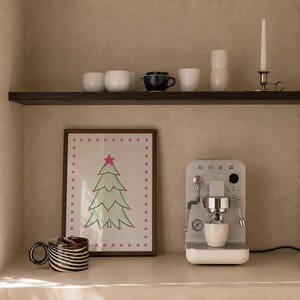 Puede incluir: Una cafetera espresso Smeg plateada con una taza blanca, un cuadro enmarcado con un &aacute;rbol de Navidad verde con estrellas rosas y una taza con patr&oacute;n de remolinos marrones y blancos. Un estante superior contiene tazas blancas, una taza azul oscuro y una vela.