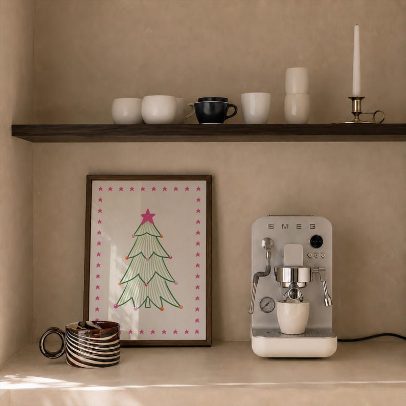 Puede incluir: Una cafetera espresso Smeg plateada con una taza blanca, un cuadro enmarcado con un &aacute;rbol de Navidad verde con estrellas rosas y una taza con patr&oacute;n de remolinos marrones y blancos. Un estante superior contiene tazas blancas, una taza azul oscuro y una vela.