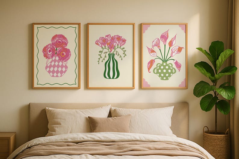 May include: Three framed floral art prints. The prints feature pink flowers in decorative vases. The vases have unique designs, including a diamond pattern, vertical stripes, and polka dots. The frames are a light wood colour, and the art is displayed above a bed.