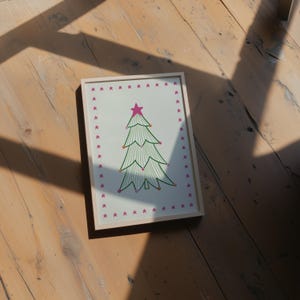 Puede incluir: Una impresi&oacute;n rectangular enmarcada que presenta un &aacute;rbol de Navidad estilizado de l&iacute;neas verdes con una estrella rosa en la punta y peque&ntilde;os puntos de colores. El &aacute;rbol est&aacute; centrado sobre un fondo blanco con un borde de estrellas rosas.