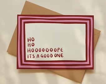 May include: A rectangular greeting card with a cream-coloured centre and a border of alternating dark red and light pink stripes. The text in dark red reads: HO HO HOOOOOOOPE IT'S A GOOD ONE. The card rests on a tan envelope.