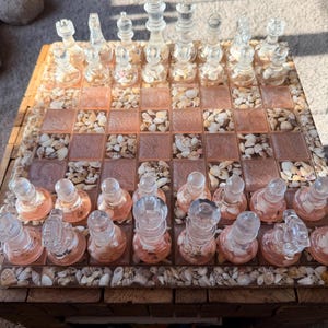 May include: A clear glass chess set with a seashell-embedded board. The chess pieces are transparent and detailed. The board has a mosaic design with pink squares and seashell accents. The wooden frame adds a rustic touch to this unique chess set.