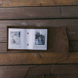 K&ouml;nnte beinhalten: Ein wei&szlig;es Lederfotoalbum mit zwei Sofortbildern darin. Das Album ist ge&ouml;ffnet und die Fotos sind sichtbar. Das Album liegt auf einer Holzoberfl&auml;che.