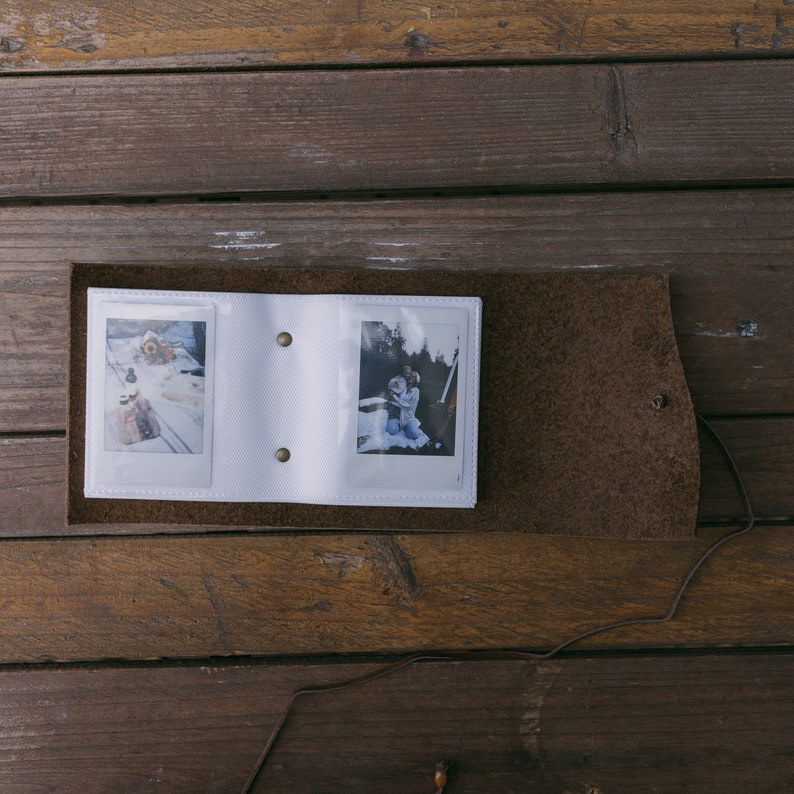 K&ouml;nnte beinhalten: Ein wei&szlig;es Lederfotoalbum mit zwei Sofortbildern darin. Das Album ist ge&ouml;ffnet und die Fotos sind sichtbar. Das Album liegt auf einer Holzoberfl&auml;che.