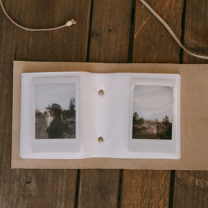 K&ouml;nnte beinhalten: Ein wei&szlig;es Lederfotoalbum mit zwei aufgeschlagenen Seiten. Die linke Seite zeigt ein Foto einer Person mit Hut, die auf einem Feld steht. Die rechte Seite zeigt ein Foto einer Landschaft mit B&auml;umen und einem Feldweg.