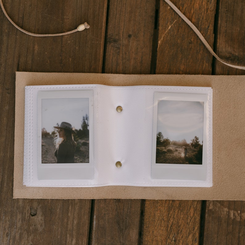 K&ouml;nnte beinhalten: Ein wei&szlig;es Lederfotoalbum mit zwei aufgeschlagenen Seiten. Die linke Seite zeigt ein Foto einer Person mit Hut, die auf einem Feld steht. Die rechte Seite zeigt ein Foto einer Landschaft mit B&auml;umen und einem Feldweg.