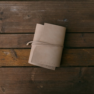 K&ouml;nnte beinhalten: Ein braunes Lederjournal mit einem braunen Lederband, das um ihn herumgebunden ist. Das Journal ist mit dem Text "LUKE + JESSICA 7.15.24" personalisiert.