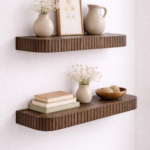 May include: Two dark brown wooden floating shelves with a grooved design are mounted on a white wall. The top shelf holds a framed floral print, a vase with white flowers, and a ceramic pitcher. The bottom shelf displays books, a vase, and a wooden bowl.