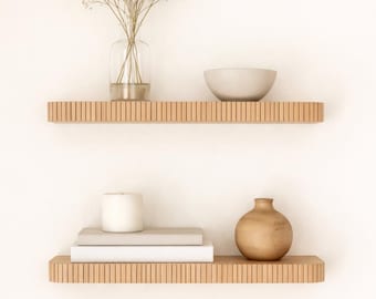 May include: Two wooden floating shelves with various decorative items. The top shelf holds a glass vase with dried flowers and a gray bowl. The bottom shelf displays a stack of books, a white candle, and a wooden vase. The shelves are made of light-colored wood.