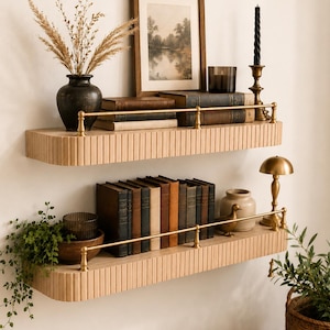 May include: Two light-colored wooden wall shelves with vertical fluted detailing and brass railings. The shelves hold various items including books, a dark vase with dried grass, a framed landscape print, a black candle, and a small brass lamp.