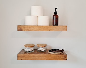 May include: Two wooden shelves with a brown finish. The top shelf has a stack of four white toilet paper rolls and a brown glass bottle with a black pump dispenser. The bottom shelf has two clear glass jars with wooden lids and a brown leather headband.