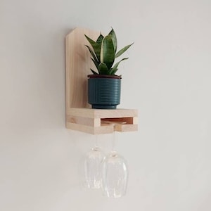 Può includere: Un ripiano a muro in legno con una piccola pianta in vaso e due bicchieri da vino appesi sotto. Il ripiano è in legno chiaro e ha un design semplice.