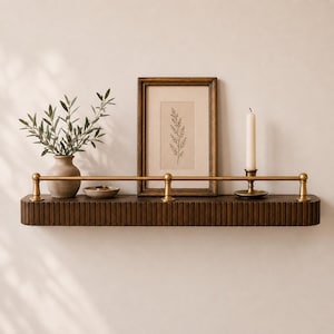 May include: A dark brown wooden wall shelf with a vertical fluted texture and a horizontal brass rail. The shelf holds a small beige vase with green leaves, a small bowl, a framed botanical print, and a white taper candle in a brass holder.