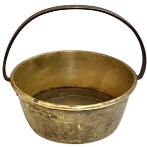 May include: A vintage brass cauldron with a dark metal handle. The cauldron has a rounded bowl shape and a slightly tarnished finish, suggesting age and use. The handle arches over the top, providing a way to carry or hang the pot.