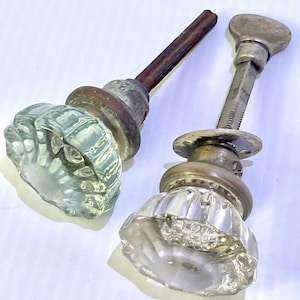 May include: Two antique glass door knobs. One knob has a clear, faceted glass design with a rusty brown metal stem. The other knob has a similar clear glass design with a silver metal stem.