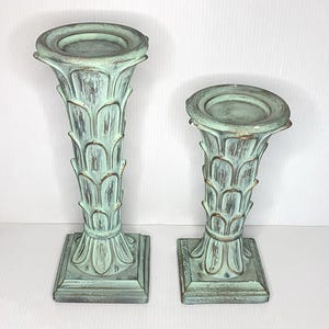 May include: Two pillar candle holders with a distressed mint green finish and gold-toned accents. Each holder features a flared top, a column body with a repeating scalloped leaf pattern, and a square base. The set includes one taller and one shorter piece.