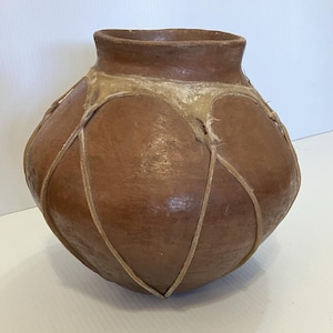 May include: A brown, rounded earthenware pot with a narrow neck. The pot is adorned with a light-colored, woven pattern that wraps around its body. The pot has a rustic, handmade appearance.