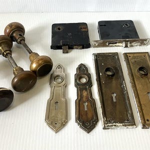May include: An assortment of antique door hardware, including several doorknobs in brass and dark brown, along with black and brass-toned lock sets and decorative keyhole covers. The hardware shows signs of age.