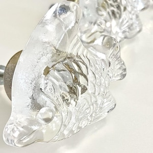 Könnte beinhalten: Schrankknöpfe aus klarem Glas in Form von Fischen. Die Knöpfe haben ein detailliertes Fischschuppenmuster und sind auf einer Metallbasis montiert. Geeignet für Möbel oder Dekorationsprojekte.
