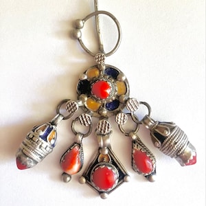 May include: A silver-toned metal brooch featuring a circular top with a pin mechanism and five dangling pendants. The central circular element and the pendants are inlaid with red, yellow, and dark blue stones, accented by intricate metalwork details.