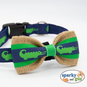 May include: A navy blue dog collar with a green alligator pattern. The collar has a matching bow tie made of burlap and green ribbon. The bow tie is adorned with a green alligator design. The collar has a black plastic buckle.