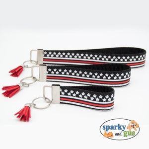 May include: Three black and white keychains with red stripes and white stars. Each keychain has a red tassel. The keychains are arranged in order of size, with the largest keychain at the top and the smallest keychain at the bottom.