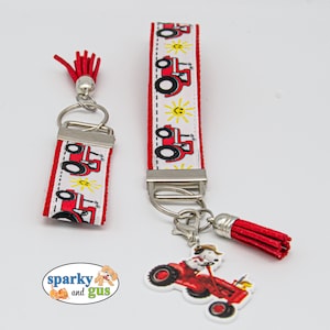 May include: Two red and white tractor themed keychains with red tassels. The larger keychain has a silver ring and a silver clasp. The smaller keychain has a silver ring and a silver loop. The smaller keychain has the text "sparky and gus" on the bottom.