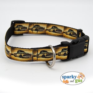 May include: A black dog collar with a yellow and brown patterned ribbon featuring a vintage car design. The collar has a silver metal D-ring and a black plastic buckle.