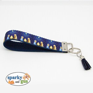 May include: A navy blue wristlet keychain with a repeating pattern of a nativity scene and fireworks. The keychain has a silver-colored metal clasp and a navy blue tassel. The brand logo "sparky and gus" is in the lower left corner.