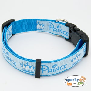 May include: A light blue dog collar with the word "PRINCE" printed in light blue, along with a crown graphic. The collar has a black plastic buckle and a metal D-ring. The collar is designed for a dog.