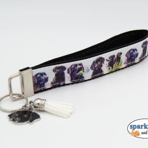 May include: A black and white wristlet key fob featuring a repeating pattern of Labrador Retrievers. The key fob has a black strap and a silver-tone metal clasp, key ring, and a dog-shaped charm. A white tassel hangs from the key ring.