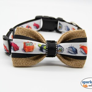 May include: A brown burlap bow tie with a white ribbon featuring a colorful sushi print. The bow tie is attached to a black dog collar.