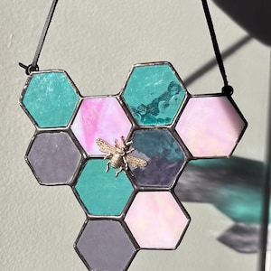 Peut inclure: Vitrail en forme de nid d'abeille avec un accent d'abeille dorée. Le design hexagonal présente des nuances de turquoise, de rose et de violet. Un cordon noir est attaché pour l'accrocher. Idéal pour la décoration intérieure.