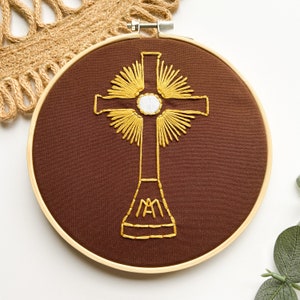 May include: A brown embroidery hoop with a gold embroidered cross with a white center circle and the letters "MA" at the base.
