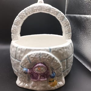 May include: White ceramic snowman igloo shaped bowl with a handle. The snowman is wearing a purple coat and blue mittens and is holding a broom.