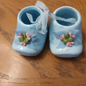 May include: A pair of blue ceramic baby shoes with a light blue ribbon and embroidered flowers. The shoes are tied together with a light blue ribbon.