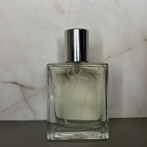 May include: A clear, rectangular perfume bottle with a silver cap. The bottle is filled with a light-colored liquid. The background is a marble-patterned wall.