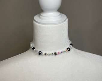May include: A white beaded necklace with black and colourful letter beads spelling out "SELF CARE".