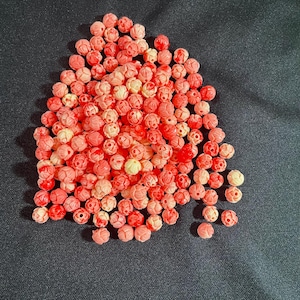 May include: A collection of small, round beads in shades of pink and red, scattered on a dark background. The beads have a carved floral design, with some showing a lighter, almost white color.