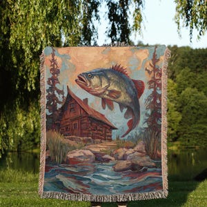 May include: A woven tapestry blanket featuring a detailed painting of a fish leaping above a cabin by a river. The artwork includes shades of blue, brown, and red. The blanket has a fringed edge.