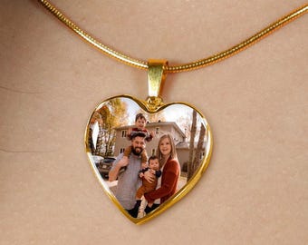May include: Gold heart-shaped necklace with a photo of a family of four. The photo shows a man, a woman, and two children. The man is holding the older child on his shoulders, and the woman is holding the younger child in her arms. The family is smiling and looking at the camera.