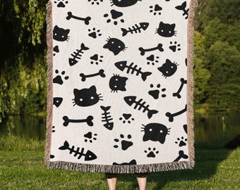 May include: A cream-colored throw blanket with a repeating pattern of black cat faces, paw prints, fish bones, and dog bones. The blanket has a fringed edge and is held up in front of a person standing on grass.