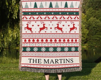 May include: A woven Christmas blanket with a festive pattern of red reindeer, trees, and snowflakes on a cream background. The blanket has a green and red border and the text "THE MARTINS" at the bottom.