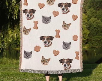 May include: Cream-colored throw blanket featuring repeating images of dogs, cats, paw prints, hearts, and bone shapes. The blanket has a fringed edge and is held up by a person standing on green grass.