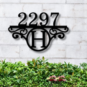 May include: Black metal house number sign with the number 2297 and the letter H in a circle. The sign is mounted on a white brick wall with green plants in front of it.